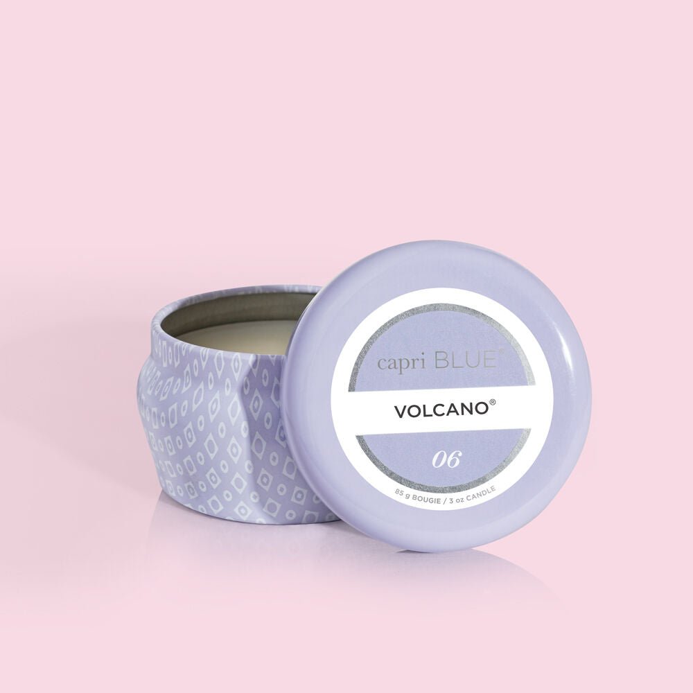 A light purple Capri BLUE Volcano Aqua Mini Tin Candle with a patterned lid resting against the base lies open on a pink background. The label reads Capri BLUE VOLCANO 06, revealing the unlit candle inside, hinting at tropical fruit and sugared citrus notes.