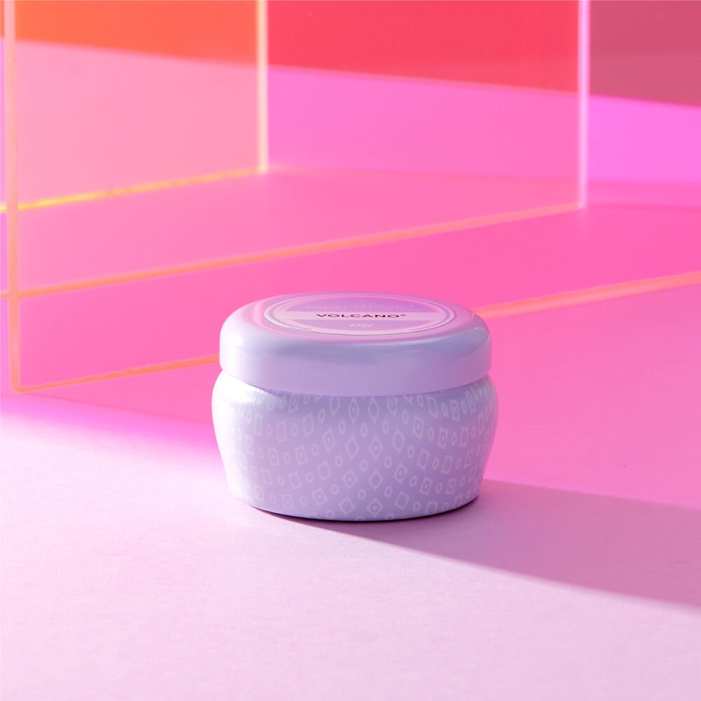 A small, round lavender-colored container labeled Capri BLUE - Volcano 3oz Aqua Mini Tin Candle rests on a pink surface. The background has pink and orange transparent panels, casting sugared citrus hues that enhance the ambiance with a warm glow.