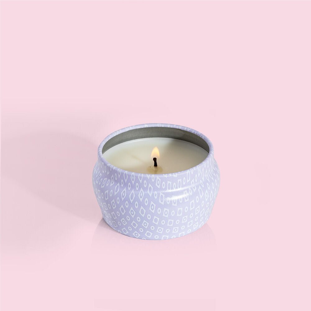 A Capri BLUE - Volcano 3oz Aqua Mini Tin Candle in a patterned tin emits sugared citrus notes. Its flame casts a soft glow against the solid pastel pink backdrop.