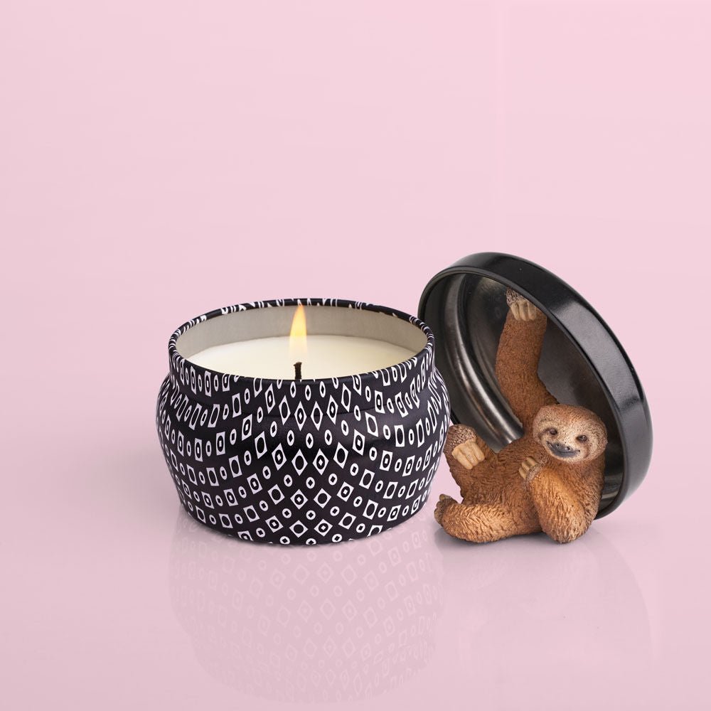 The Capri BLUE - Volcano 3oz Black Mini Tin Candle, placed on a pink surface, features its lid off to show a small sloth figurine on the underside. The scent of sugared citrus fills the air around its black and white patterned holder.