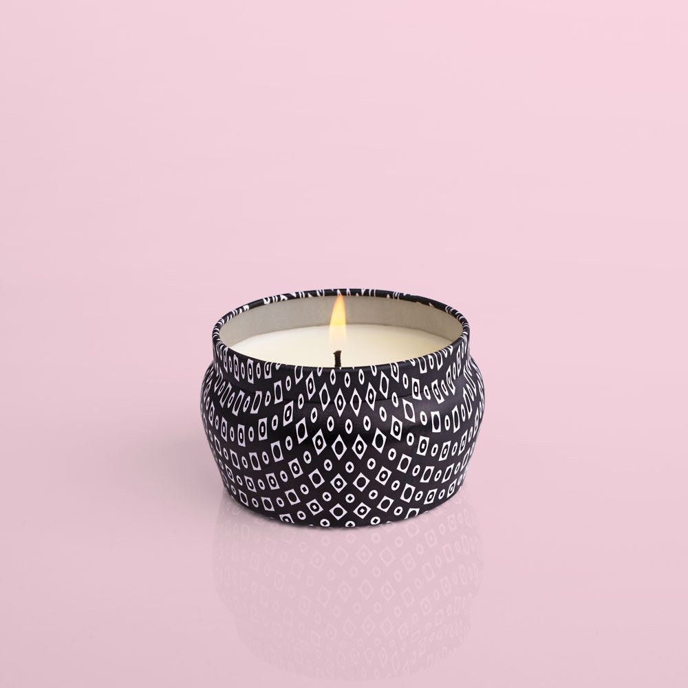 The Capri BLUE - Volcano 3oz Black Mini Tin Candle sits in a round metal tin with a black and white diamond pattern, releasing sugared citrus notes. The setup includes a solid pastel pink background.