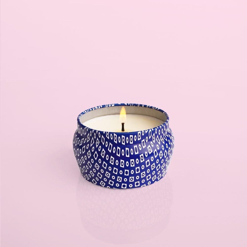 A Capri BLUE - Volcano 3oz Blue Mini Tin Candle, known for its iconic scent, is housed in a round decorative tin with a blue and white geometric pattern against a pale pink background.