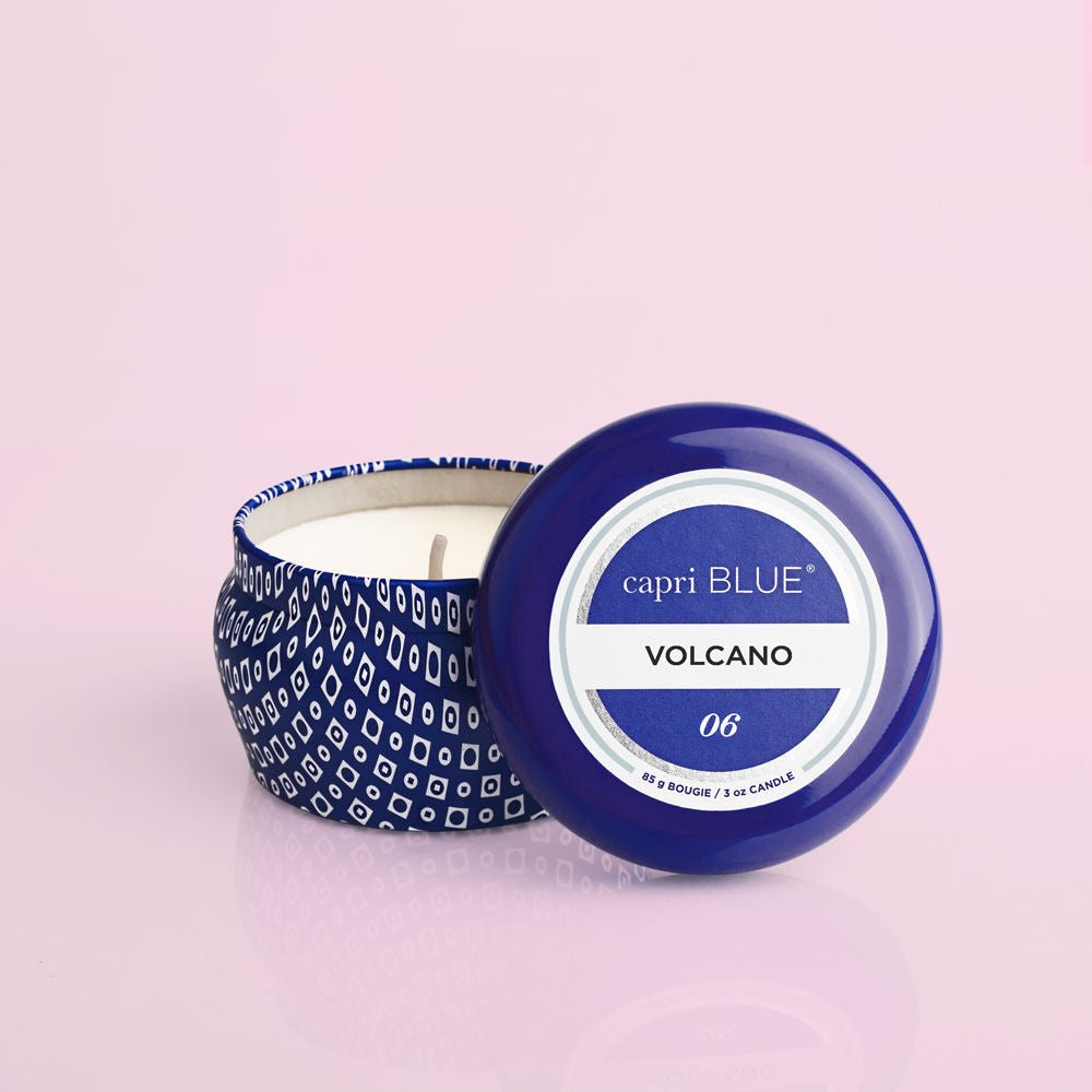 A Capri BLUE - Volcano 3oz Blue Mini Tin Candle, known for its iconic scent perfect for getaways, rests open on a soft pink surface with the lid beside it, revealing a white candle inside.