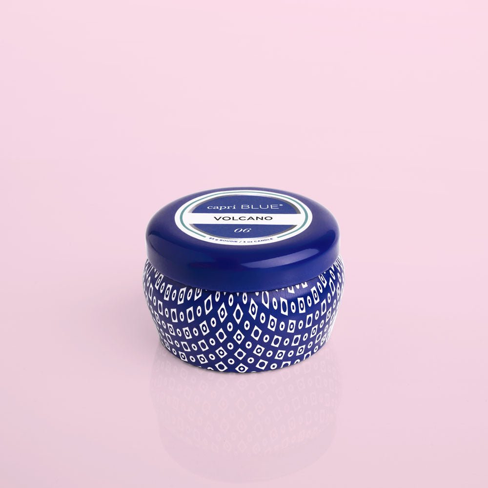 On a pink backdrop sits a blue and white patterned candle jar with a round lid. The label says Capri BLUE - Volcano. This iconic scent hints at dreamy escapes, and with the Capri BLUE - Volcano 3oz Blue Mini Tin Candle, you can carry this fragrance wherever you roam.