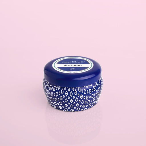 On a pink backdrop sits a blue and white patterned candle jar with a round lid. The label says Capri BLUE - Volcano. This iconic scent hints at dreamy escapes, and with the Capri BLUE - Volcano 3oz Blue Mini Tin Candle, you can carry this fragrance wherever you roam.