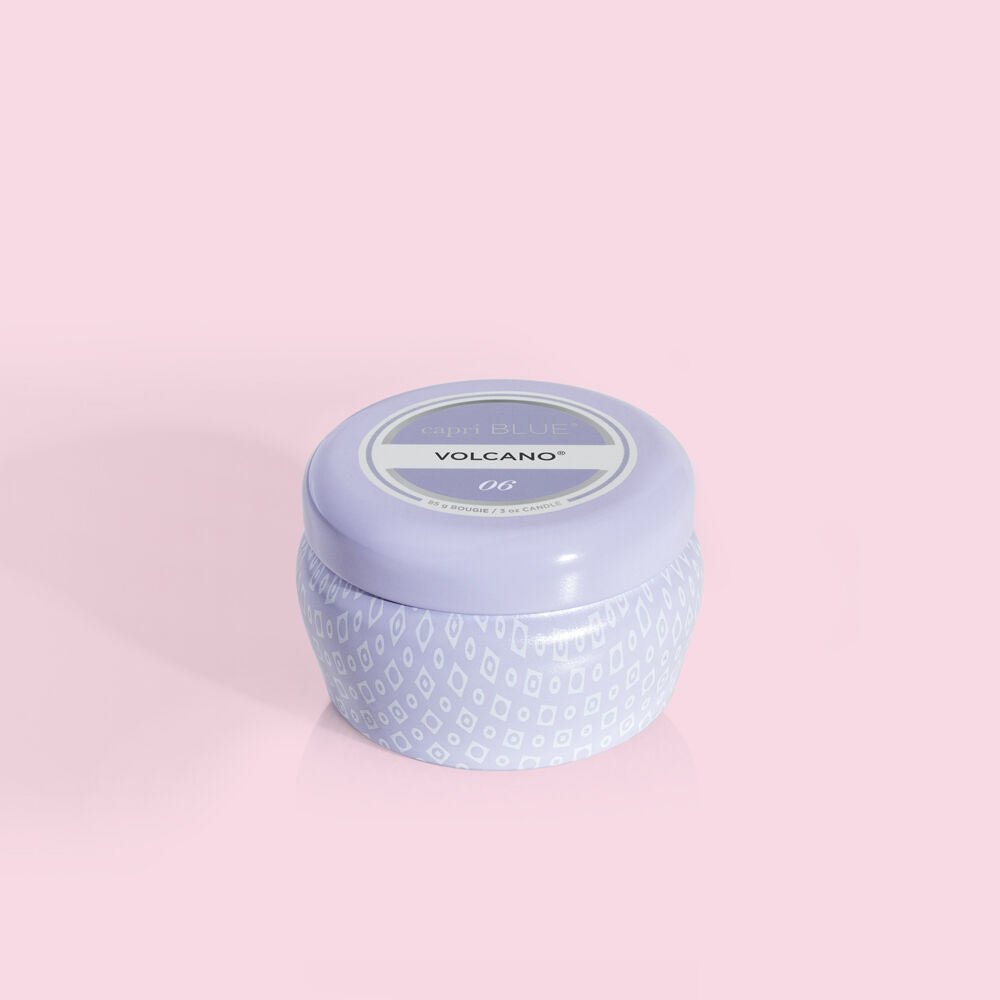 A Capri BLUE Volcano 3oz Lavender Mini Tin Candle, featuring a patterned base and a lid labeled Capri Blue Volcano OG on a soft pink background, emits the lively aroma of tropical fruits and sugared citrus.