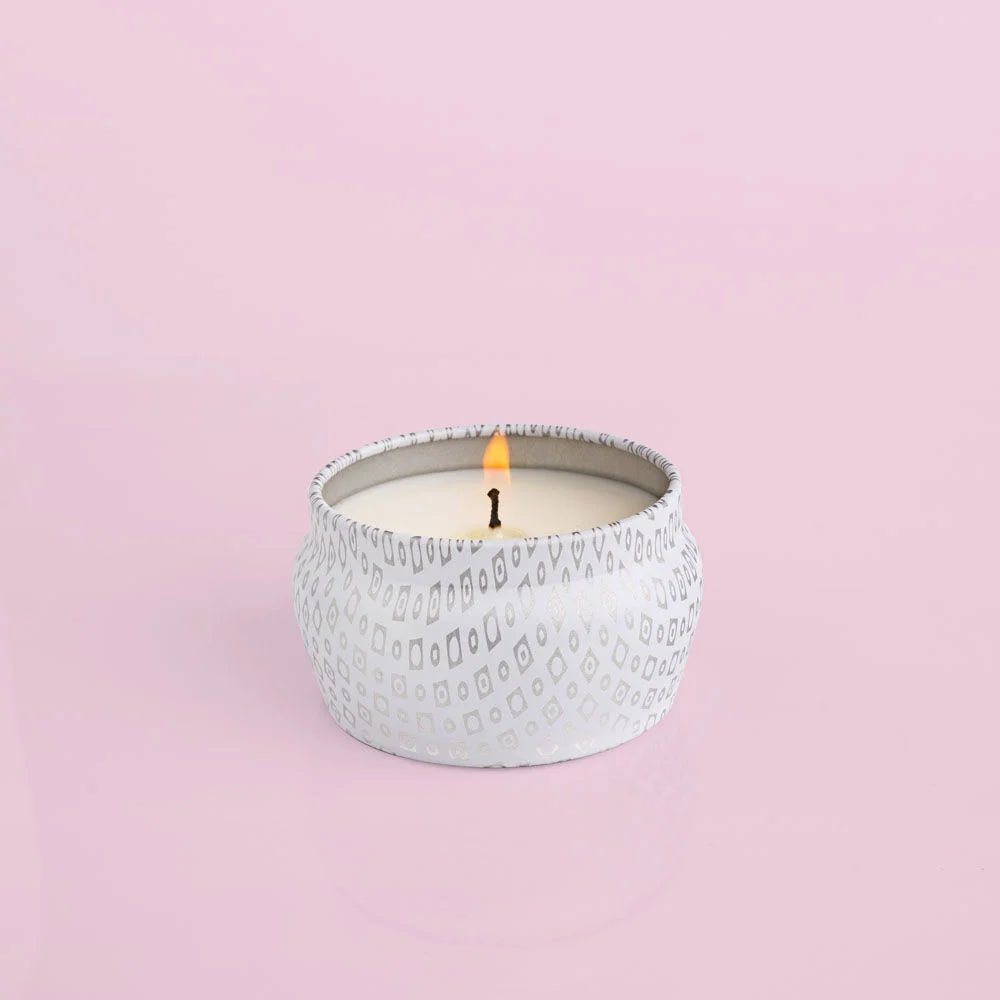 A Capri BLUE Volcano 3oz White Mini Tin Candle, featuring a decorative geometric design, sits on a light pink surface, releasing its subtle fragrance. The flame is small and centered.