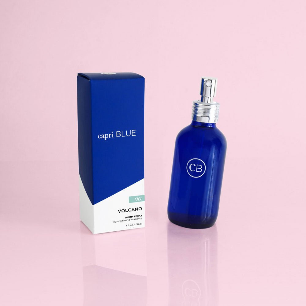 A blue bottle of Capri BLUE - Volcano 4oz Room Spray with a silver top stands next to its matching box labeled Capri BLUE, emitting a tropical citrus-sugary scent, all beautifully set against a pale pink background.