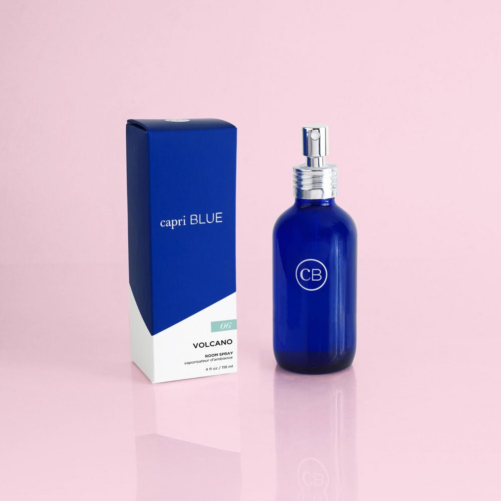 A Capri BLUE Volcano 4oz Room Spray in a blue glass bottle sits elegantly next to its matching box labeled Capri BLUE VOLCANO Body Spray, offering a tropical fragrance of citrus and sugary notes, stylishly placed on a pale pink surface.