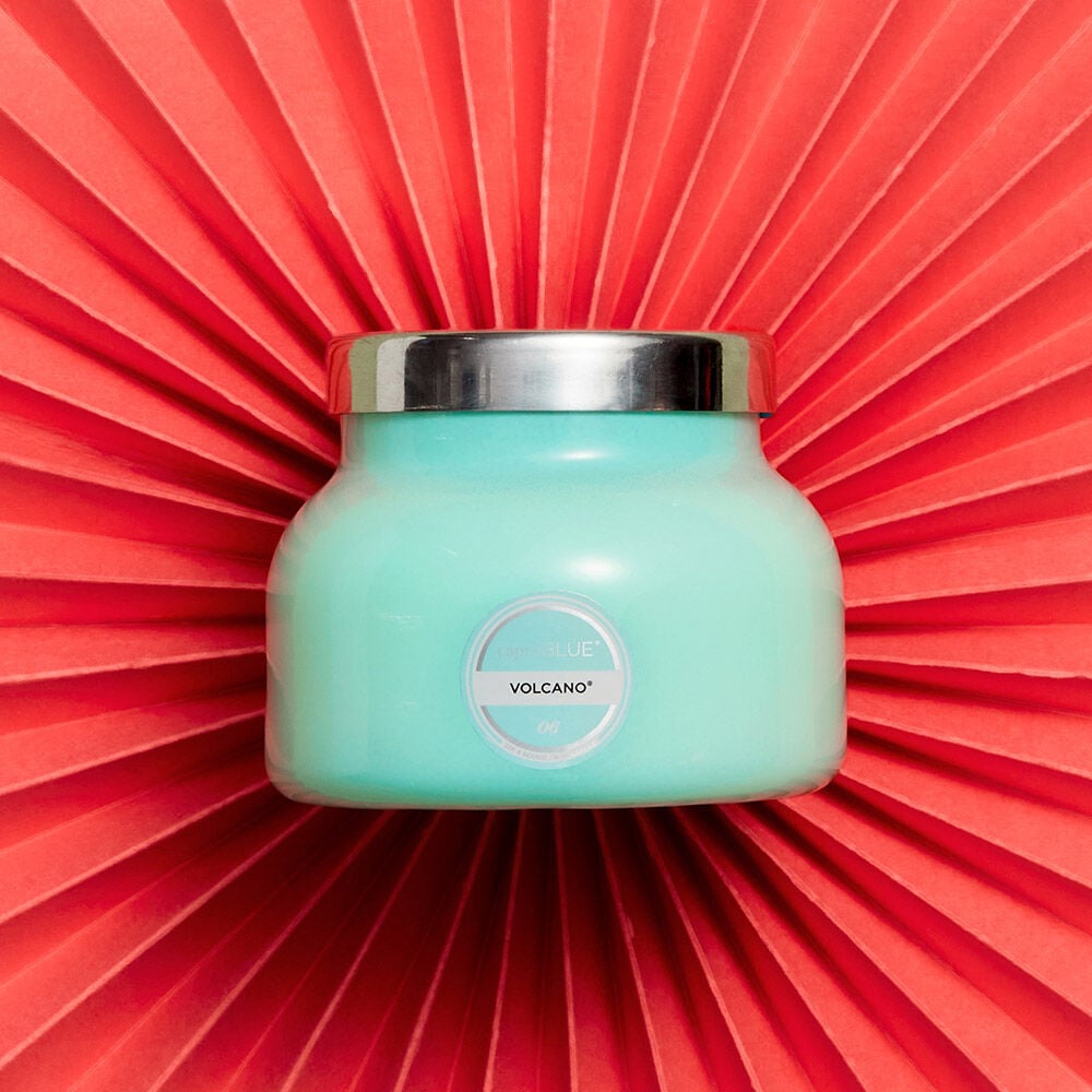 The Capri BLUE - Volcano 8oz Aqua Petite Candle, with a stylish silver lid, stands out against a bright coral background. Its sleek design and enticing Volcano fragrance, featuring sugared citrus notes, are perfectly highlighted by the vibrant contrast.