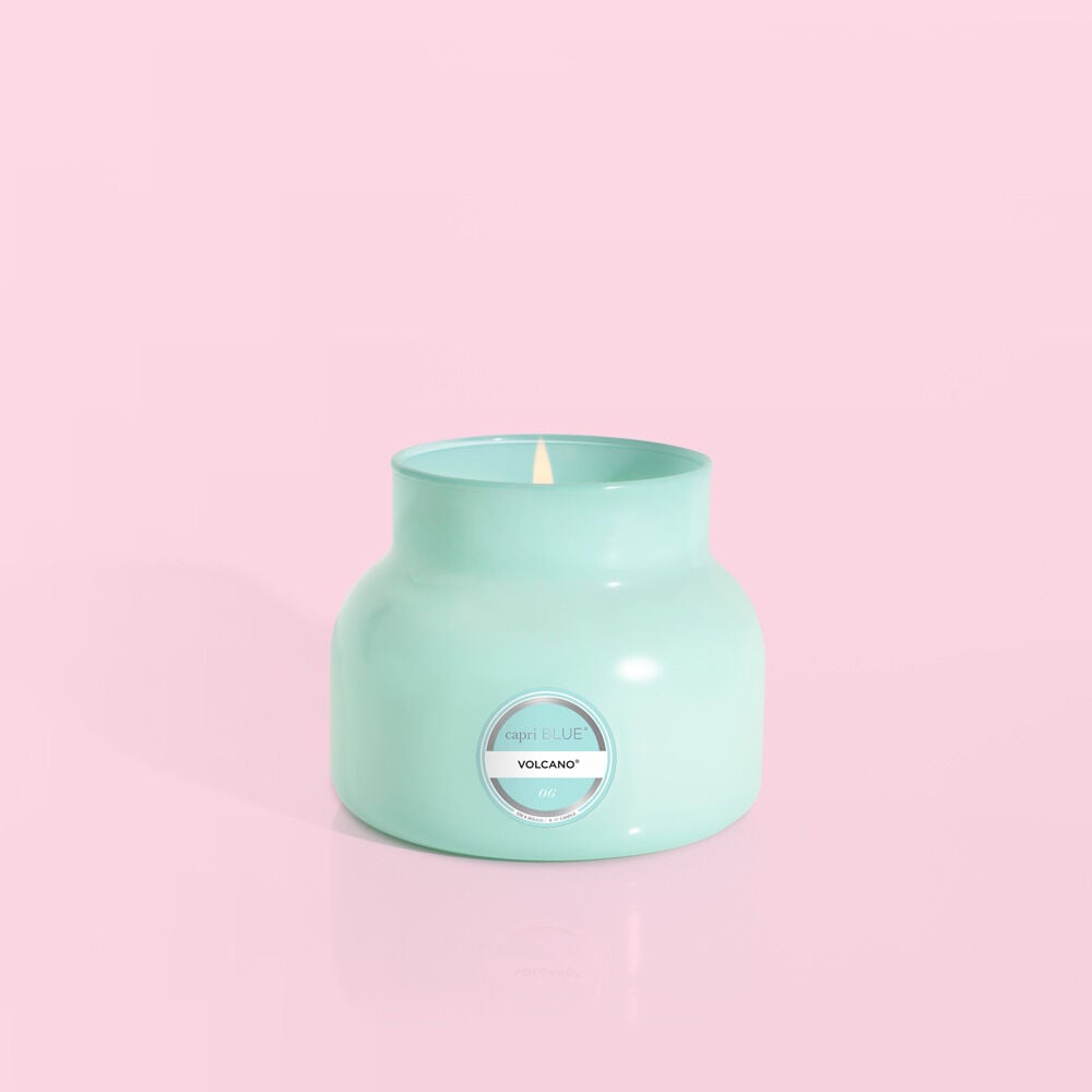 An aqua jar candle from Capri BLUE, labeled Volcano, is lit against a pale pink background, evoking the fragrant allure of tropical fruits and sugared citrus.