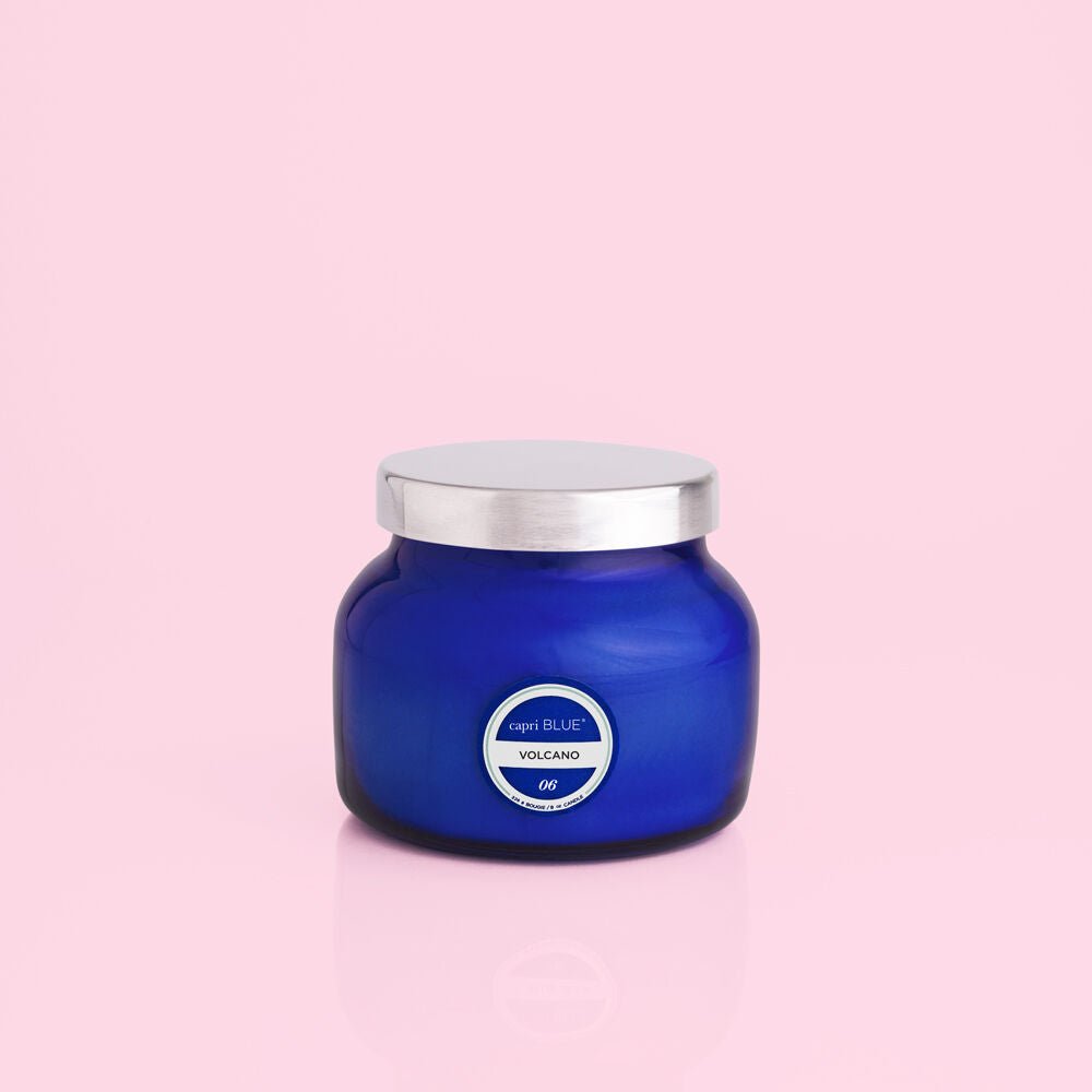 The Capri BLUE - Volcano 8oz Blue Petite Candle features sugared citrus notes, comes in a cobalt blue jar with a silver lid, and is shown against a solid light pink background.