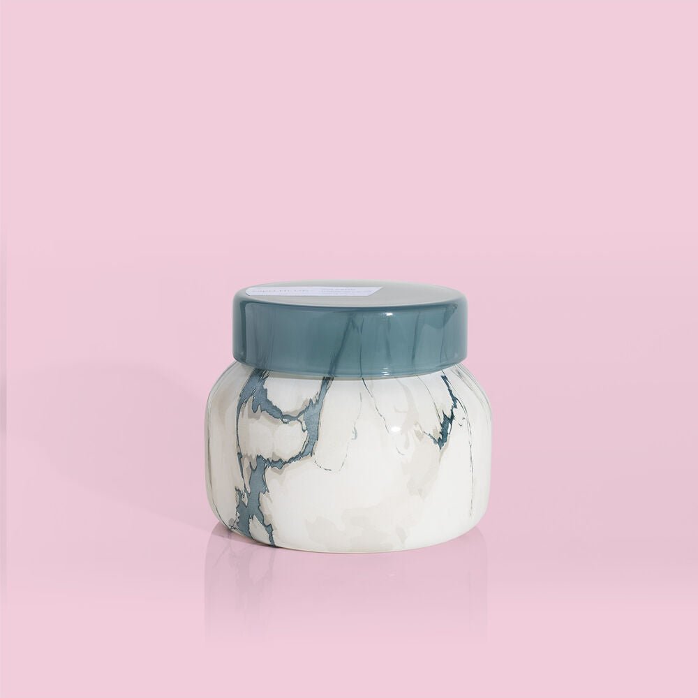 A Capri BLUE Volcano 8oz Marble Petite Candle with a dark gray lid sits centered on a pale pink background. This rounded, minimalist candle adds an elegant touch—ideal for enjoying the signature Volcano fragrance by Capri BLUE.