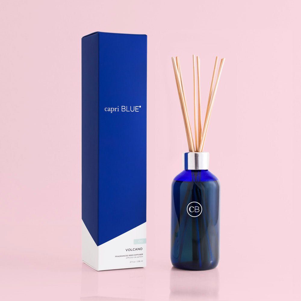 The Capri BLUE - Volcano 8oz Reed Diffuser exudes modern elegance against a pink background, with a dark blue bottle and wooden reeds topped with a silver cap, standing next to its blue box and adorned with a circular CB logo.