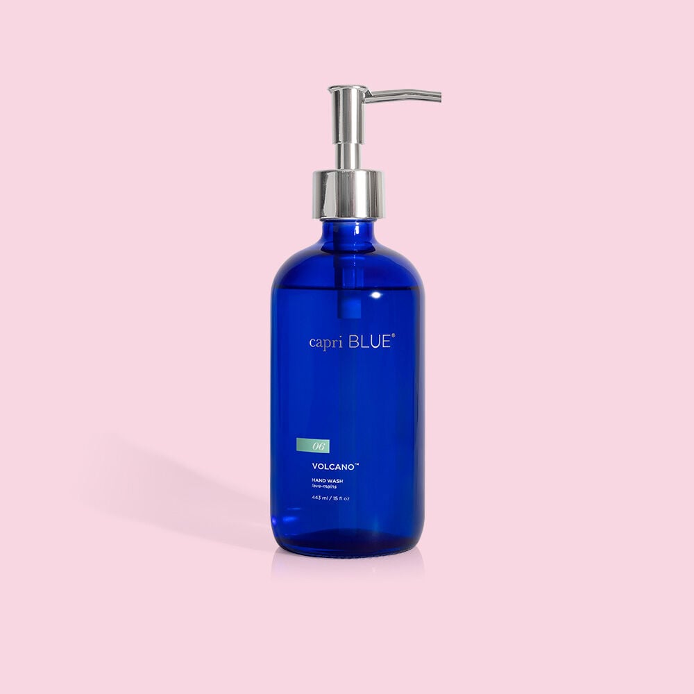 A sleek blue glass pump bottle, labeled Capri BLUE, displays the Volcano Hand Wash 15oz with plant-based glycerin against a pastel pink background. Featuring a silver pump dispenser, its reusable design adds an eco-friendly touch to your daily routine.
.