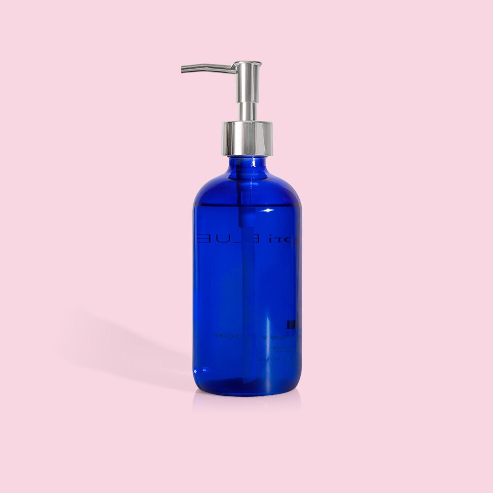 The Capri BLUE - Volcano Hand Wash 15oz in a cobalt blue glass bottle with a silver dispenser is set against a solid pink background. It features plant-based glycerin and an eco-friendly, reusable design.