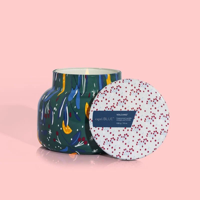 The Capri BLUE Volcano Holiday Pattern Play Petite Jar 19 oz Candle sits on a pink background, featuring colorful abstract designs on its green jar. The white lid has red speckles and a blue Capri Blue Volcano label, exuding a vibrant ambiance with its tropical fruit fragrance.