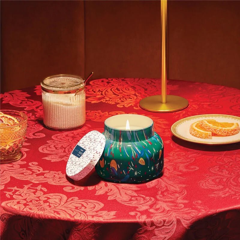 A Capri BLUE Volcano Holiday Pattern Play Petite Jar candle with a galaxy design sits on a red floral tablecloth. Beside it are its lid, a jar with another lid, tropical fruits on a plate, and a gold lamp. The warm lighting and Volcano fragrance from Capri BLUE offer a cozy ambiance.