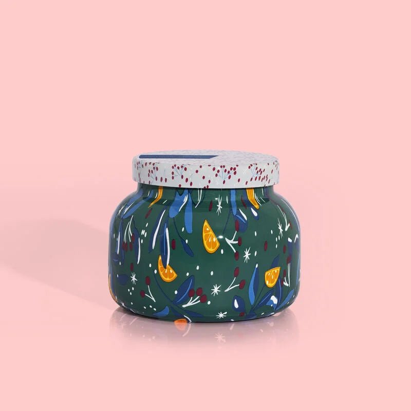 The Capri BLUE - Volcano Holiday Pattern Play Petite Jar 19 oz Candle features a speckled top, dark green body with colorful floral accents, and a vibrant design of tropical fruits and orange slices on a pink background.