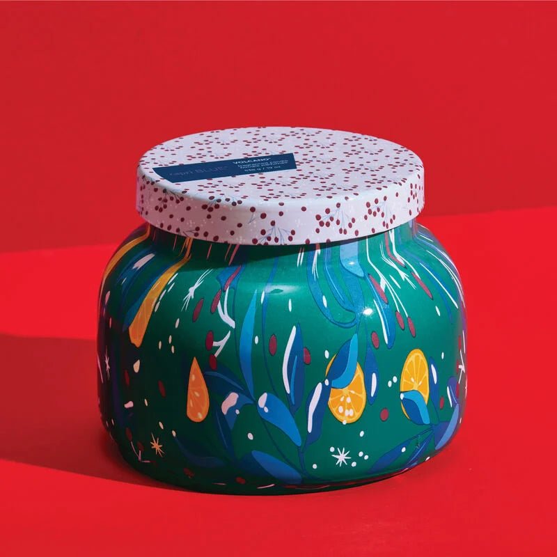 The Capri BLUE - Volcano Holiday Pattern Play Petite Jar Candle is a 19 oz green jar featuring abstract plant and tropical fruit patterns in yellow, orange, and white. It has a festive holiday design with a vibrant red background and is topped with a white lid adorned with red speckles.