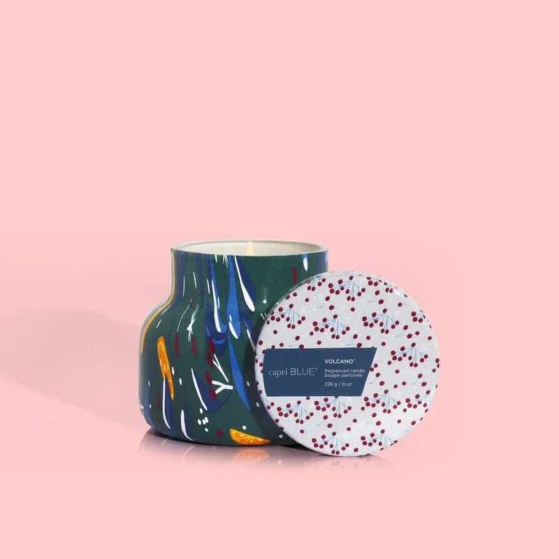 The Capri BLUE - Volcano Holiday Pattern Play Petite Jar 8 oz Candle features a vibrant, abstract pattern. The candle is set against a soft pink background with a lid displaying red speckled text, while shadows elegantly enhance the jars decorative appeal.