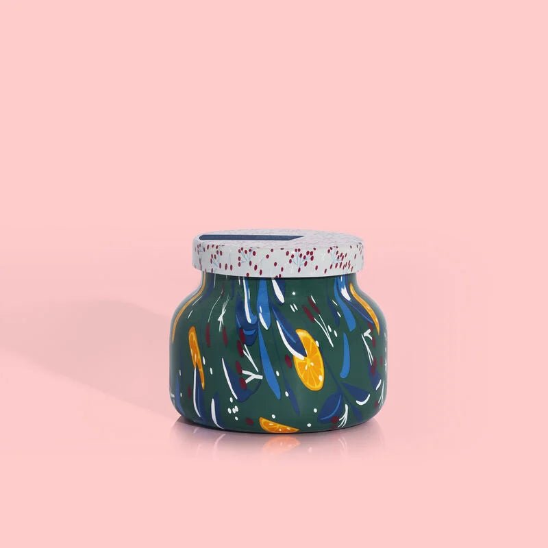 The Capri BLUE - Volcano Holiday Pattern Play Petite Jar 8 oz Candle features a round, colorful jar on a pink background, with abstract leaf-like patterns in blue, orange, and white. The lid is speckled with red dots and infused with sugared citrus undertones.