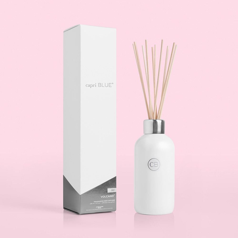 The Capri BLUE - Volcano White 8oz Reed Diffuser features CB initials on a white bottle with thin reeds, accompanied by a tall rectangular box labeled capri BLUE on a pink background. Its striking geometric design showcases the signature VOLCANO fragrance.