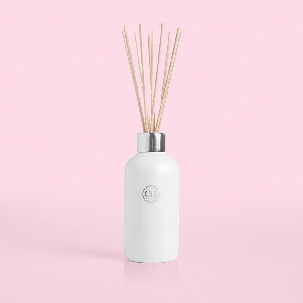 Set against a pink backdrop, the Capri BLUE - Volcano White 8oz Reed Diffuser showcases elegance with its white bottle, silver cap, and slim wooden reeds. An embossed CB circle on its front adds stylish sophistication to this home fragrance essential.