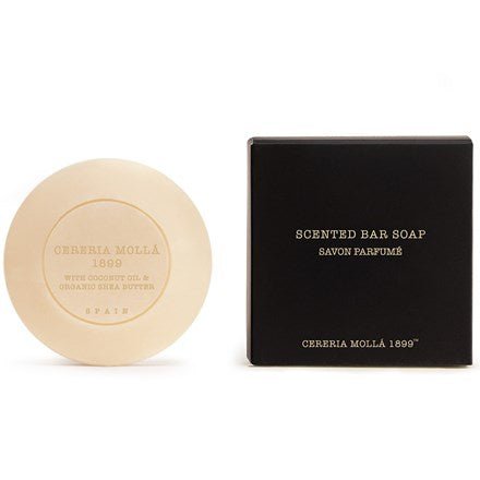 A round, beige Cereria Molla soap engraved with Cereria Mollá 1899 rests next to a black, square box labeled Scented Bar Soap in gold. Infused with coconut oil and organic shea butter, it exudes an amber and sandalwood scent.