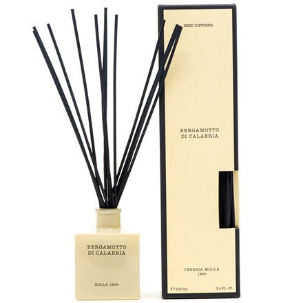 A 3.4oz beige square bottle of Cereria Molla Bergamotto di Calabria reed diffuser with black sticks is placed beside its matching rectangular box. Emitting a lush Mediterranean fragrance, the label reads Cereria Molla. Both are beautifully set against a crisp white background.