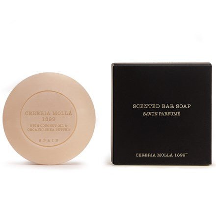 A round beige soap bar, embossed with Cereria Molla 1899 and Spain, is placed next to a black box labeled Scented Bar Soap featuring coconut oil and organic shea butter details. The air is gently infused with Bulgarian Rose from the Cereria Molla - Bulgarian Rose & Oud 3.5oz Scented Bar Soap.