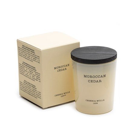 A 2.6oz Moroccan Cedar soy wax candle by Cereria Mollá is in a glass jar with a black lid, next to its beige box displaying the brand name Cereria Molla.