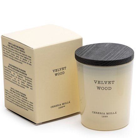 The Cereria Molla 2.6oz scented candle, Velvet Wood, infused with aromatic spices, features a black wooden lid and comes elegantly enclosed next to its matching cream-colored box.