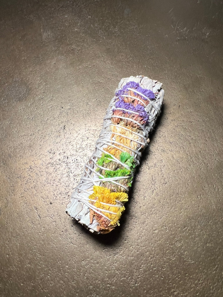 The Good Vibes Sage 4 by Designs by Deekay is wrapped in a spiral pattern with string, adorned with colorful dried flowers, perfect for smudging and inviting positive energy. It rests on a textured brown surface, showcasing vibrant colors from purple to yellow.