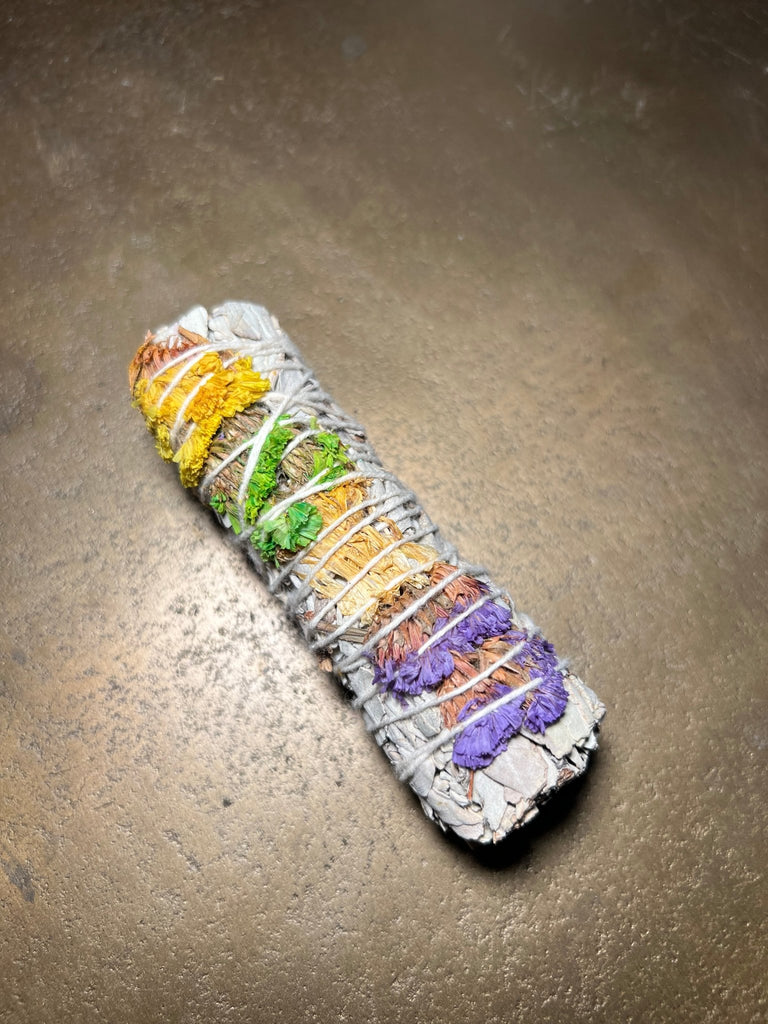 Designs by Deekays 4 Good Vibes Sage bundle, woven with vibrant yellow, green, orange, and purple dried flowers, rests on a textured brown surface. Ideal for smudge rituals to invite positive energy into your space.