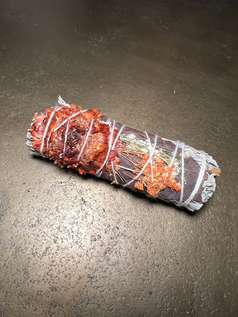 A smudge stick wrapped in string with dried orange and red flowers sits on a dark surface. This Keepsake Sage 4 by Designs by Deekay emits a fragrant blend of sage and herbs, evoking remembrance.