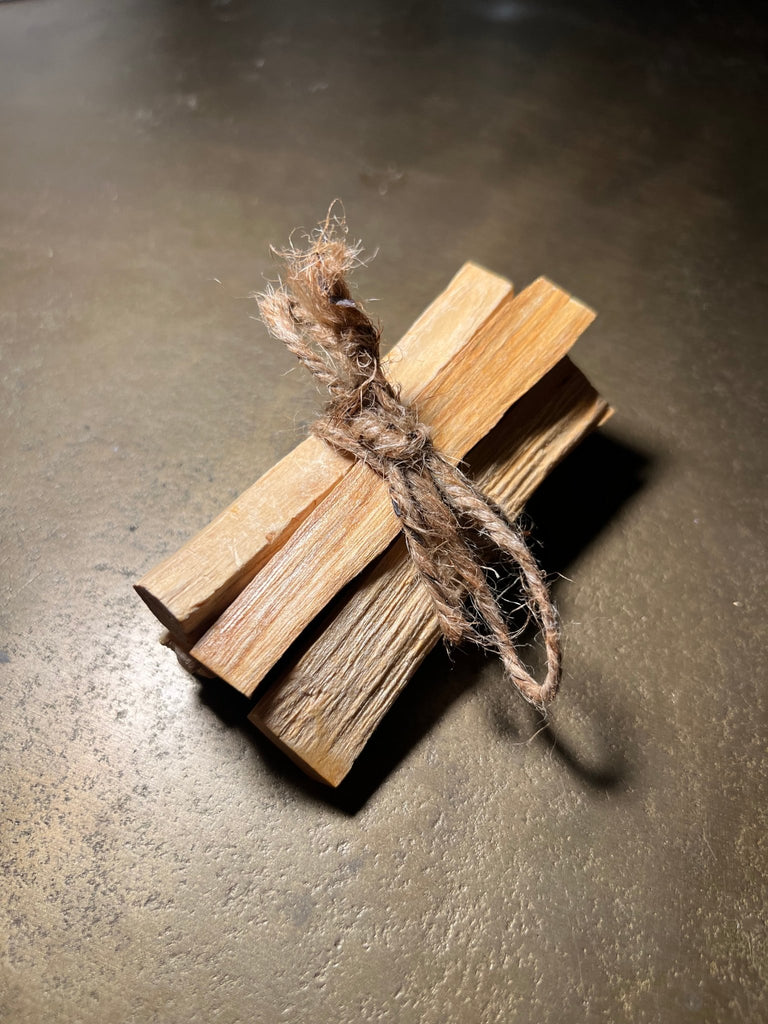 A Designs by Deekay Sage - Palo Santo Bundle of 5 sticks, bound with twine, sits on a textured brown surface. The light woods visible grain is believed to clear negative energy and attract good fortune into your space.