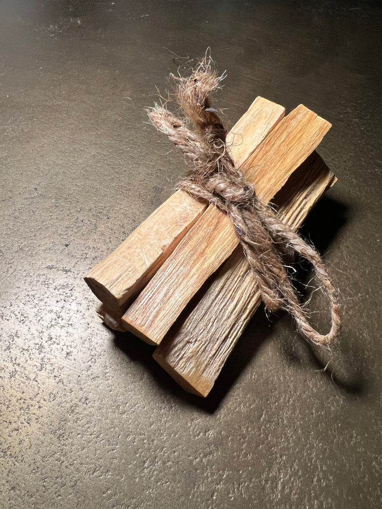 The Sage - Palo Santo Bundle by Designs by Deekay features five slightly rough, light brown palo santo sticks tied with rustic twine on a dark surface, ready to clear negative energy. The contrasting knotted and frayed twine enhances its earthy appeal.