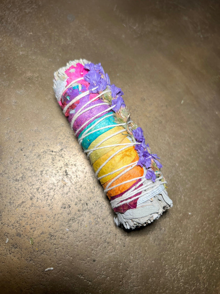 A 4 Rainbow Rose Sage bundle by Designs by Deekay lies on a brown surface, wrapped with twine. This sage, symbolizing love and attraction, is adorned with pink, purple, and yellow dried flowers.