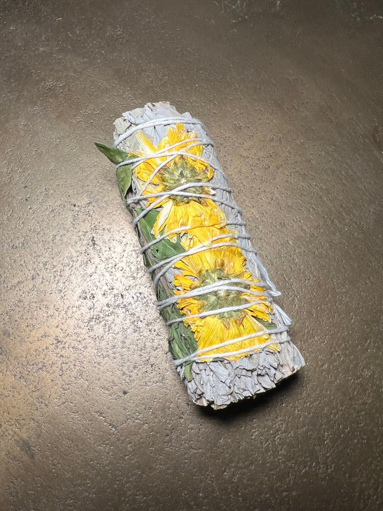 A 4 Sunflower Sage by Designs by Deekay, wrapped with twine and adorned with two sunflowers, sits on a textured brown surface. This handcrafted smudge tool is believed to bring positive energy and tranquility to any space.