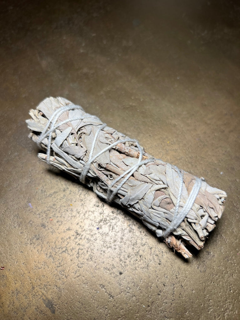 A 4 white sage bundle from Designs by Deekay rests on an earthy surface, with its light grayish-green leaves forming a cylindrical shape. Used for smudging to clear negative energy, lighting highlights the intricate details of this aromatic product.