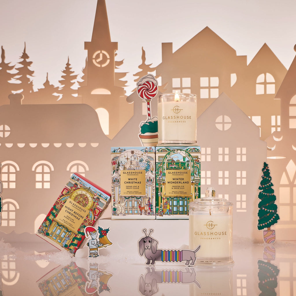 A festive holiday scene featuring the Glasshouse Fragrances 2.1oz Christmas Candle Trio, a decorated chocolate bar, and Christmas-themed paper cutouts like a dachshund, lollipop, and a snowy village backdrop.
