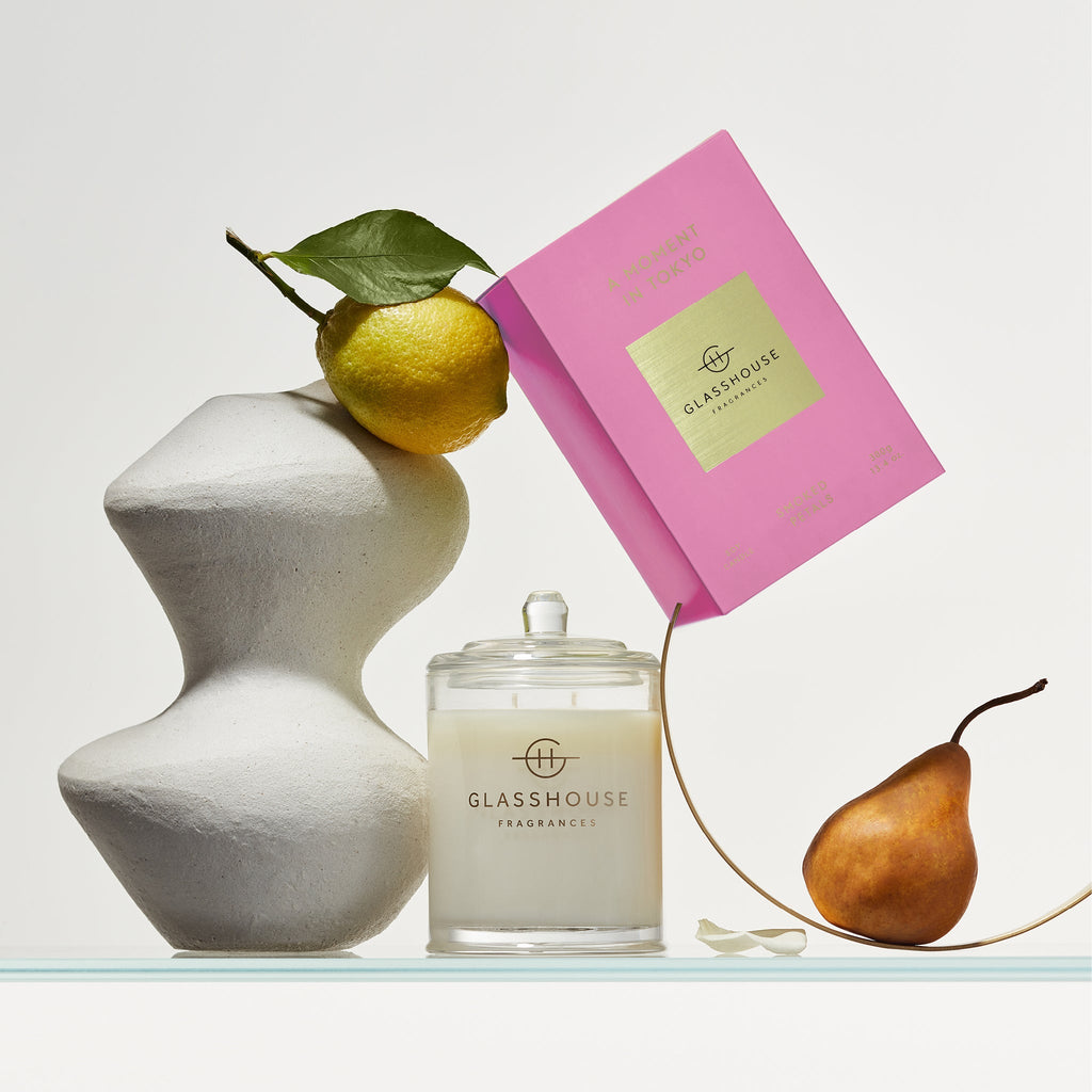 A Glasshouse Fragrances - A Moment In Tokyo 13.4oz Candle sits beside a pink box, while a sculpted white vase holding a lemon with leaves and a pear on glass add an artistic touch, highlighting the fragrances Tokyo Smoked Petals and Incense Violet Leather aroma.