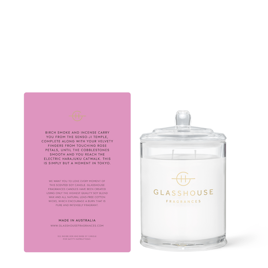 Beside a pink card revealing its scent of incense, violet, and leather, evoking Tokyos essence with smoked petals, sits a Glasshouse Fragrances A Moment In Tokyo 13.4oz triple scented candle in a clear glass jar with lid. The base reads Made in Australia.