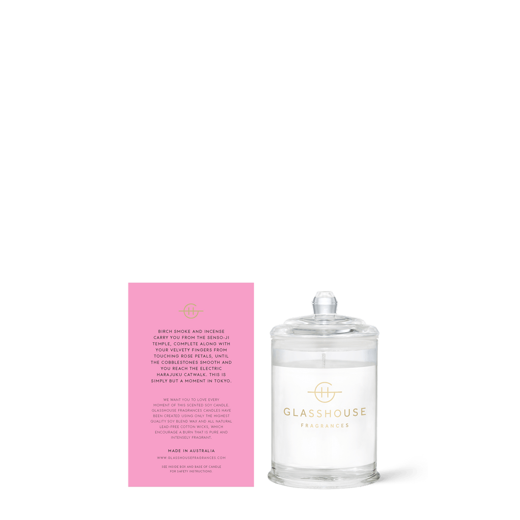 A clear jar labeled Glasshouse Fragrances - A Moment In Tokyo Mini 2.1oz Candle, likely crafted from natural soy wax, sits next to a pink card with product details and might feature the enticing aroma of Tokyo Smoked Petals.