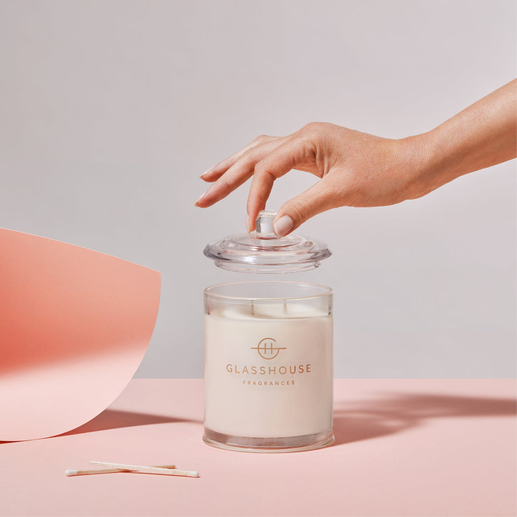 A hand lifts the lid off a Glasshouse Fragrances - Arabian Nights 13.4oz candle in a clear glass container on a pink surface with two matches. A curved sheet of pink paper enhances the backdrop, evoking hints of saffron and white oud allure.