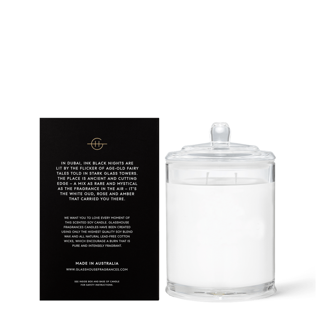 A clear glass Arabian Nights 13.4oz candle with a lid is accompanied by a black card, describing its inspiration from Dubais nights. This triple-scented soy candle by Glasshouse Fragrances is infused with saffron and white oud, showcasing its poetic essence and brand details.