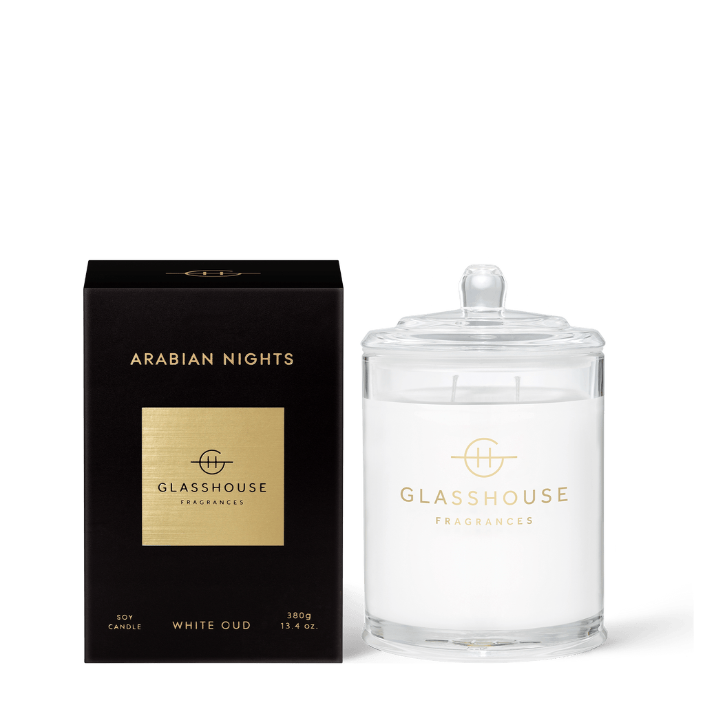 Next to a black box with gold Arabian Nights, Glasshouse Fragrances, and White Oud text stands a triple-scented soy candle in a glass jar with a lid. Infused with rich saffron and white oud, the 13.4oz Glasshouse Fragrances candle mirrors the luxurious branding.