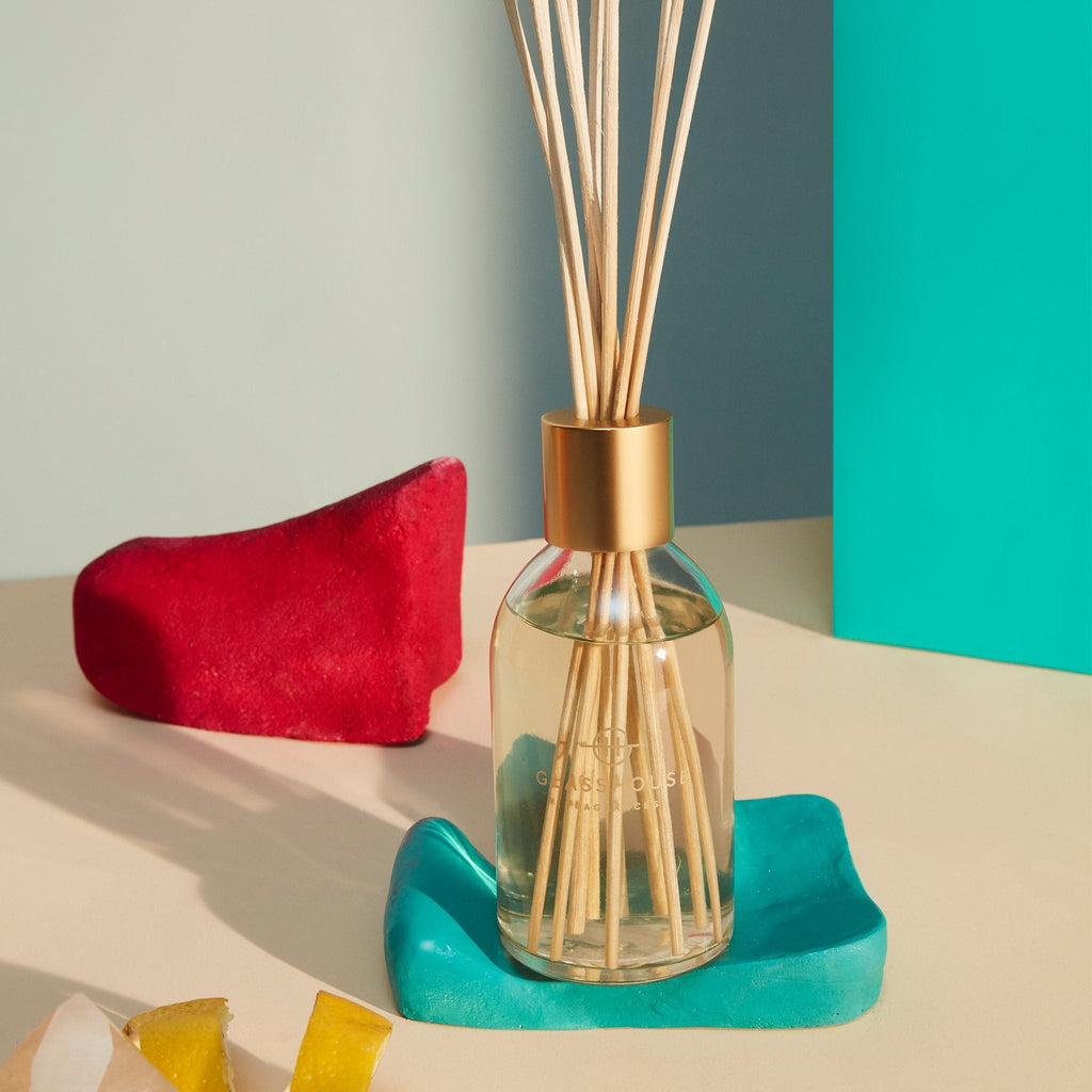 The Glasshouse Fragrances - Arabian Nights Diffuser, featuring wooden sticks in a glass design on a teal ceramic dish, is paired with a red abstract object and colored geometric shapes against a neutral background. The ambiance is enriched by the delicate aroma of saffron and white oud.