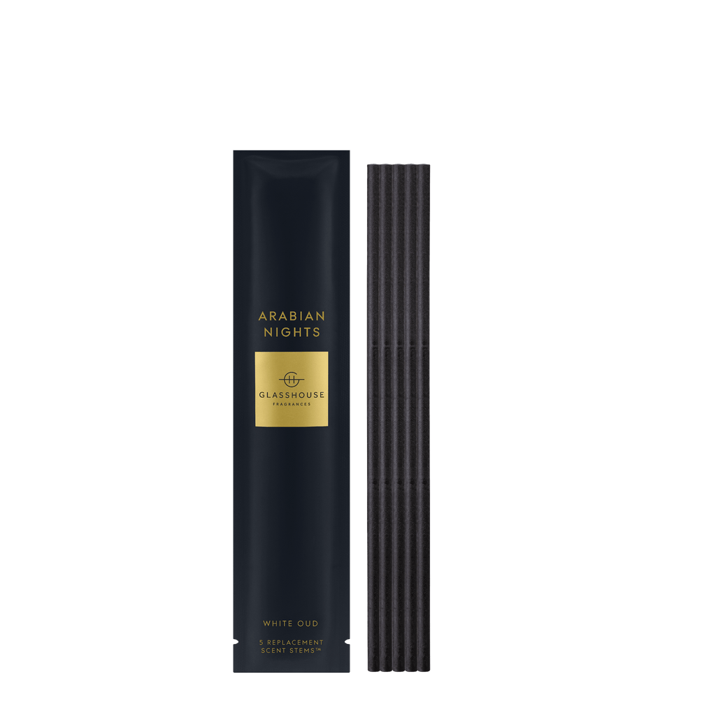 Beside a dark blue and gold box marked Glasshouse Fragrances - Arabian Nights Replacement Scent Stems, five black sticks emit a mysterious aroma. Saffron and white oud blend to create an enchanting fragrance experience by Glasshouse Fragrances.